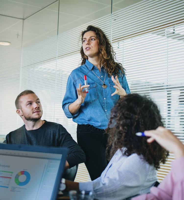 A woman in a denim shirt speaks to colleagues in a meeting room, holding a red pen, while one person listens and another takes notes on a laptop with a chart displayed.