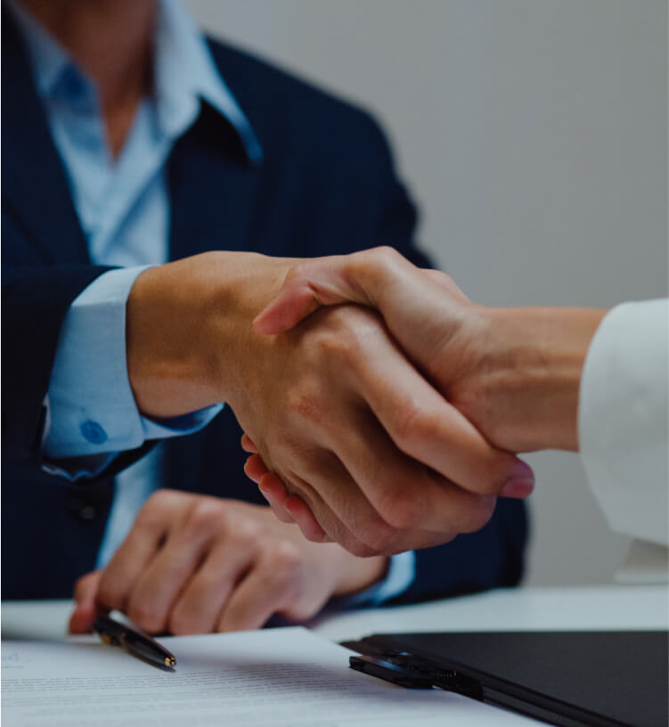 Two people in business attire shaking hands over a desk with documents and a pen visible.