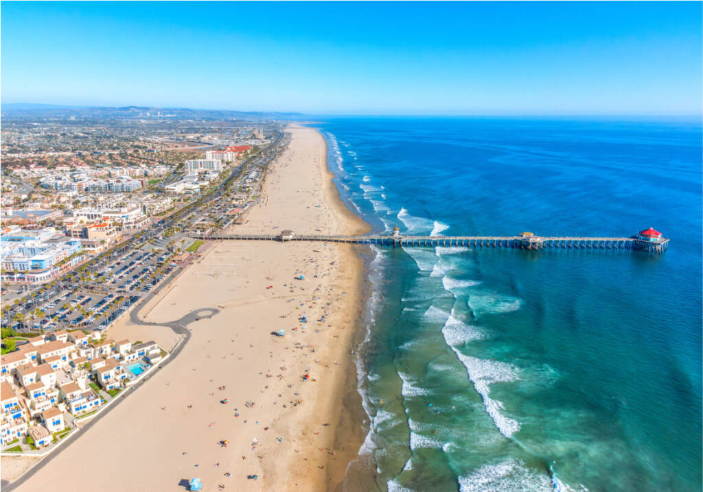 Aerial view of a long sandy beach with a pier extending into the ocean. The shoreline is lined with buildings and parked cars, while waves approach the beach.