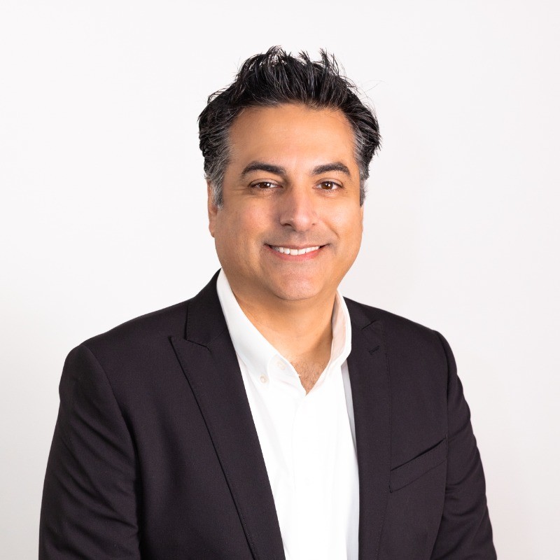 A man with dark hair wearing a black blazer and white shirt is smiling at the camera in front of a plain white background.