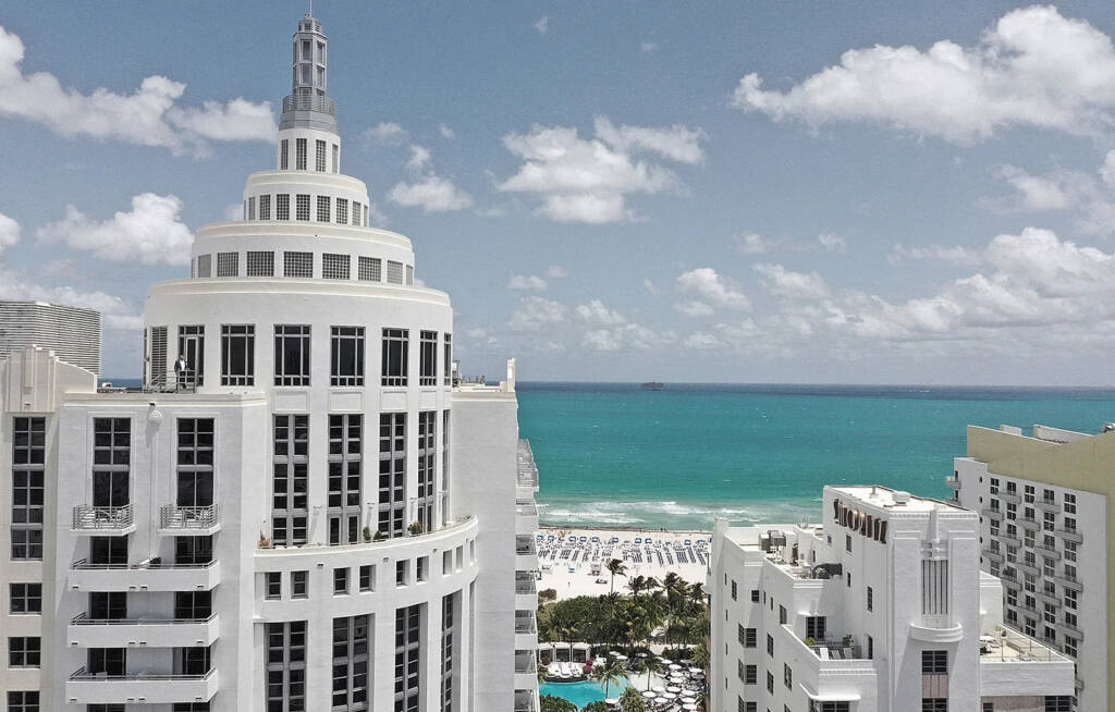 Tall white building with Art Deco design overlooking a turquoise ocean and sandy beach under a partly cloudy sky.