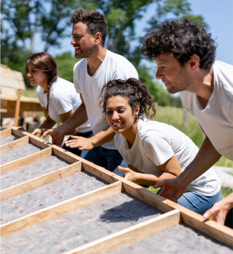 A group of people in white shirts collaboratively lift a wooden frame outdoors on a sunny day.