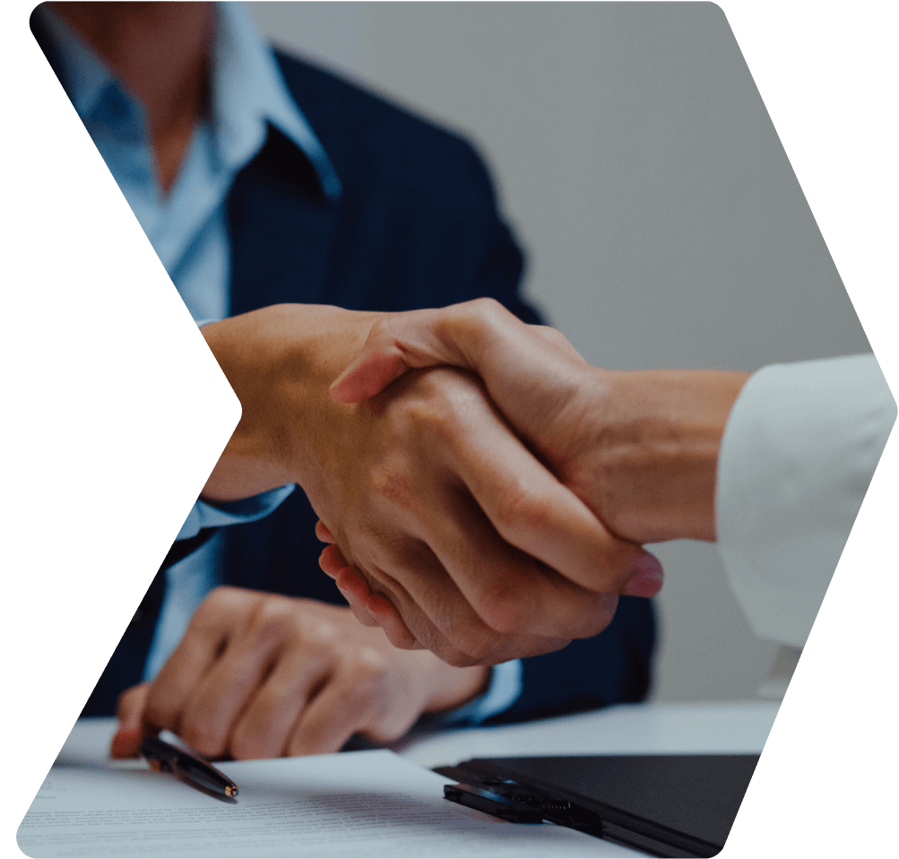 Close-up of two people shaking hands over a table with documents and a pen. One person wears a dark suit, the other a light-colored outfit.