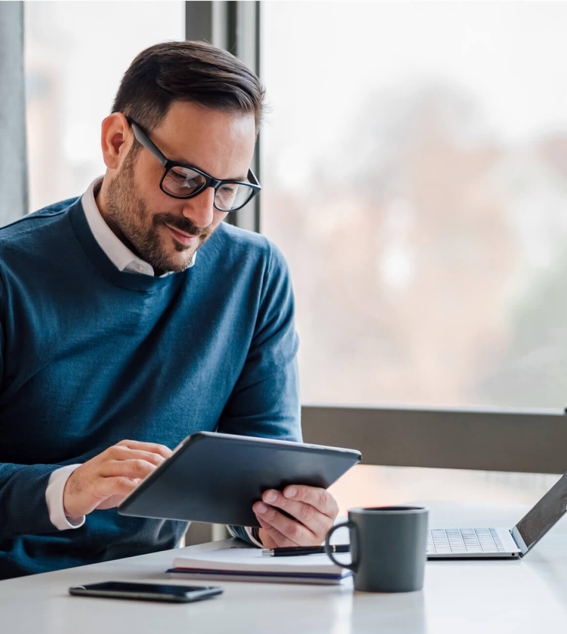 A man with glasses sits at a table using a tablet, with a laptop, coffee mug, and smartphone nearby.