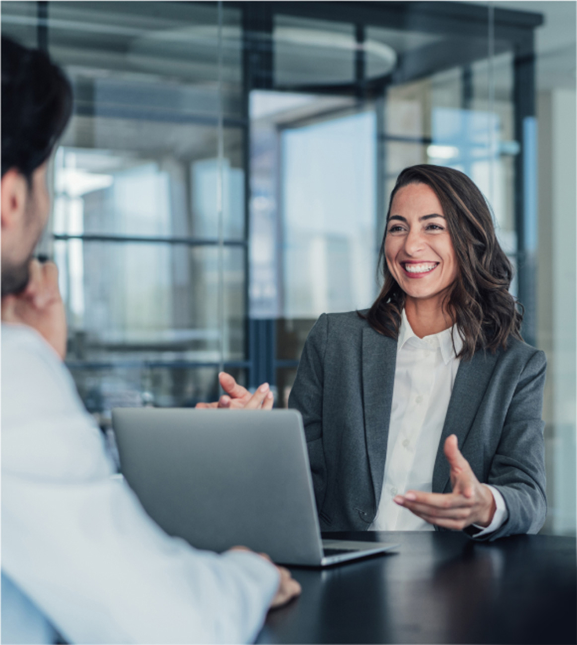 A woman in a business suit smiles and gestures while talking to a man across a table in a modern office with a laptop in front of her.
