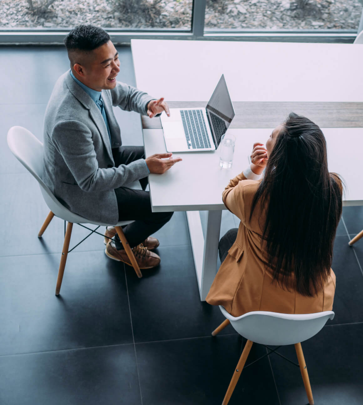 Two people sit opposite each other at a table, engaging in conversation. One person works on a laptop. They are in a modern office space with large windows.