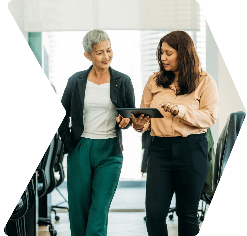 Two women walking in an office, discussing contents on a tablet.