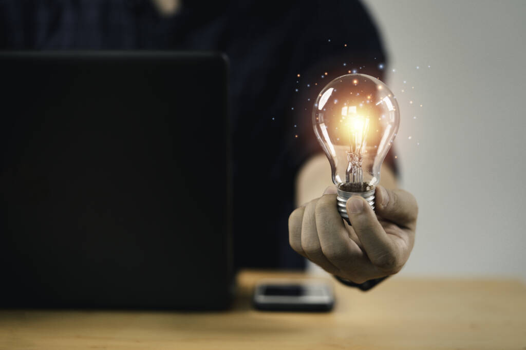 A person holds a glowing light bulb in front of a laptop on a wooden desk, symbolizing ideas or innovation.