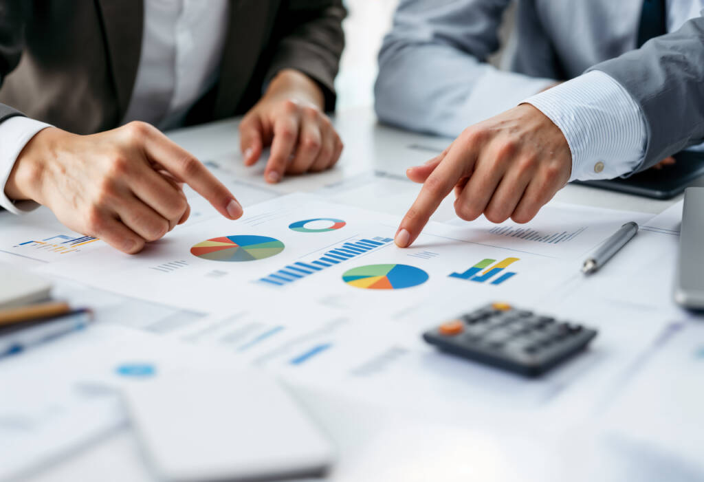 Two people review printed financial charts and graphs on a desk, pointing at data with their fingers; a calculator and documents are also visible.