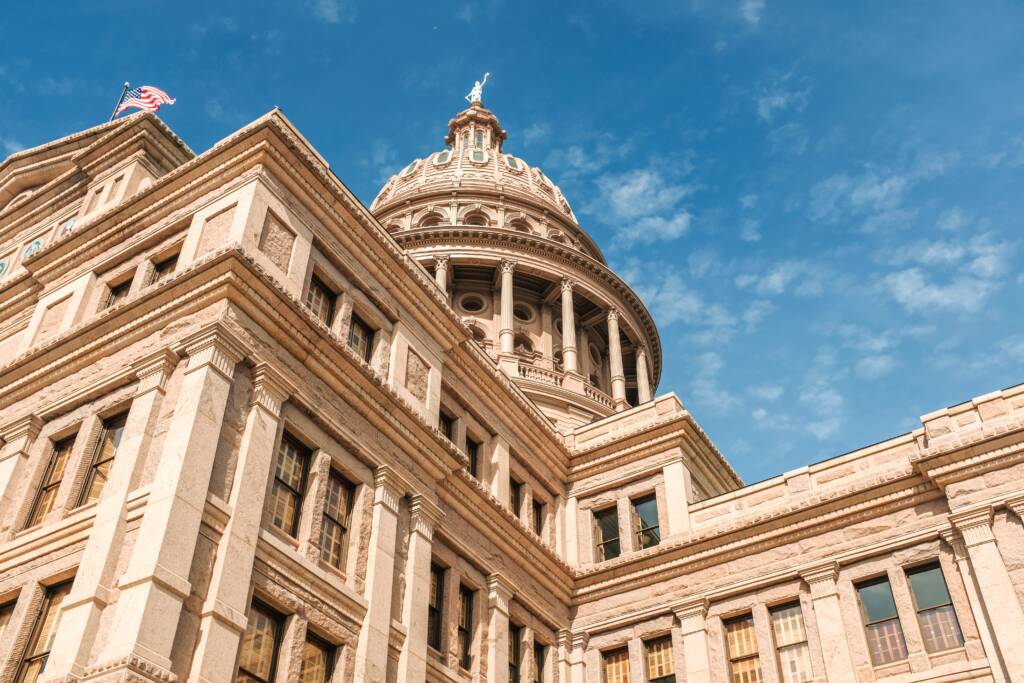 Exterior view of a large stone government building with a dome, featuring columns and an American flag, set against a blue sky with scattered clouds.