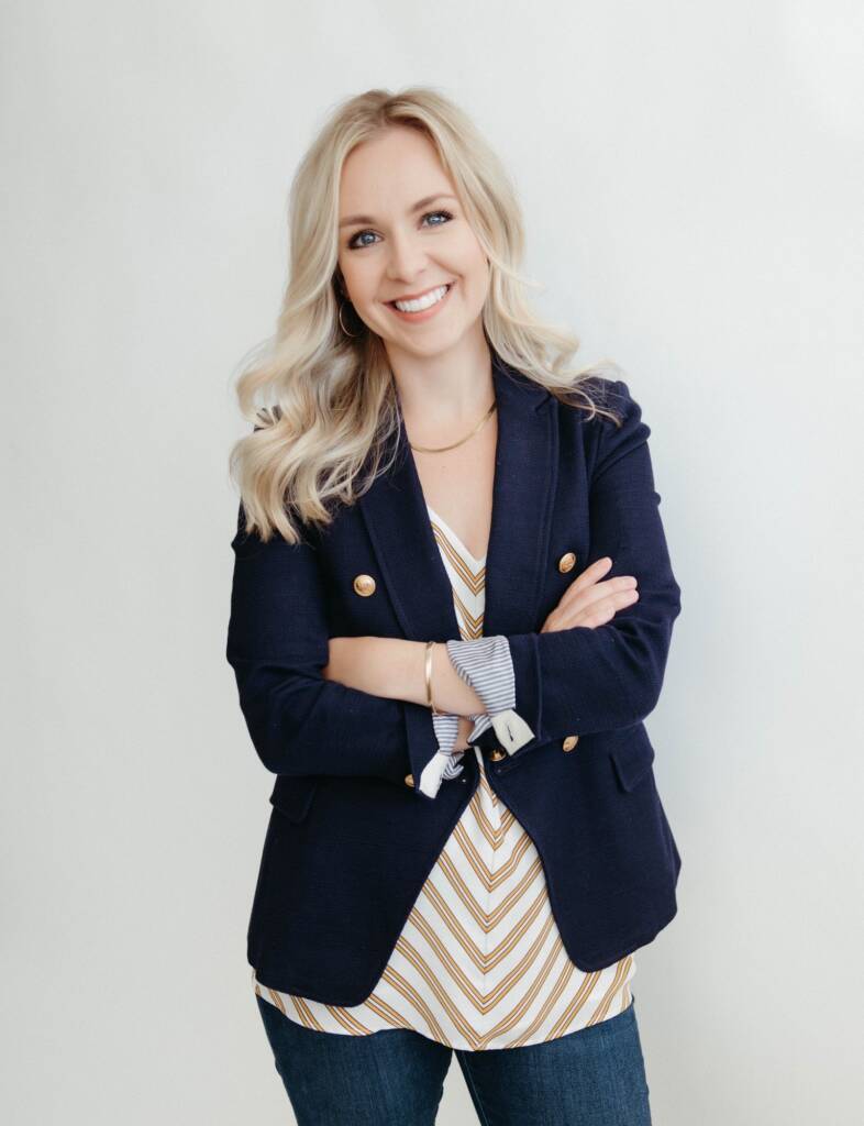 A woman with long blonde hair, wearing a navy blazer and patterned blouse, stands with arms crossed, smiling against a plain background.