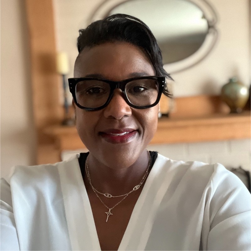 Person wearing glasses and a white blouse smiles at the camera. They are standing indoors with a wooden mantel and a round mirror in the background.