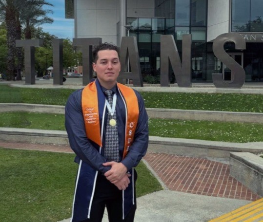 A young man wearing graduation attire stands in front of large "TITANS" letters on a campus lawn.