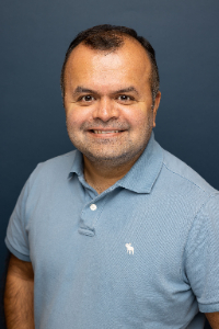 A man with short dark hair and a beard, wearing a light blue polo shirt, smiles at the camera against a plain dark blue background.