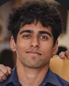 A young man with curly dark hair and a blue shirt is looking at the camera and smiling slightly.