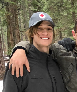 A young person wearing a gray hat with a red cross stands outdoors in a snowy forest, smiling with an arm around another person.