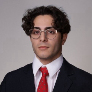 A young man with curly dark hair, wearing glasses, a white shirt, red tie, and dark suit jacket, poses for a formal headshot against a plain background.
