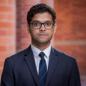 A man wearing glasses and a dark suit with a striped tie stands in front of a blurred brick background, facing the camera with a neutral expression.