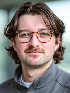 A young man with medium-length brown hair, wearing round eyeglasses and a gray jacket, looks at the camera with a slight smile.