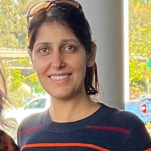 A woman with long dark hair, wearing a dark sweater and sunglasses on her head, stands outdoors and smiles at the camera. Trees and a blue truck are visible in the background.