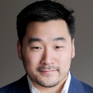 A man with short black hair, a trimmed beard, and a mustache is wearing a blue suit jacket and white shirt, posing in front of a plain gray background.