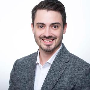 A man with short dark hair and facial hair, wearing a gray checkered blazer and white shirt, smiles at the camera against a plain white background.