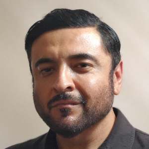 A man with short dark hair, a beard, and a mustache is facing the camera with a neutral expression. He is wearing a dark collared shirt against a plain background.