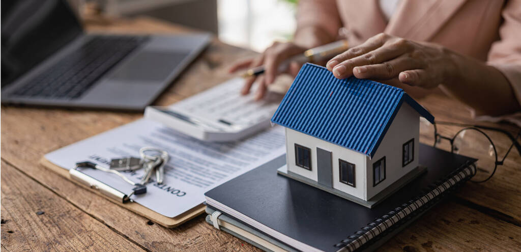 A person uses a calculator next to a contract, house model, notebook, and keys on a wooden desk.