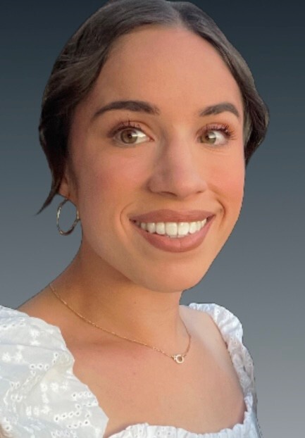 Woman with brown hair in a low bun, wearing a white top, gold hoop earrings, and a gold necklace, smiling against a gray gradient background.