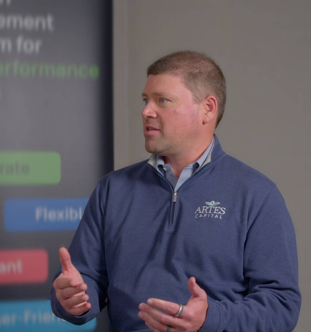 A man in a blue zip-up jacket sits and gestures while speaking. A potted plant and a black tumbler are on the table, with a blurred company sign in the background.