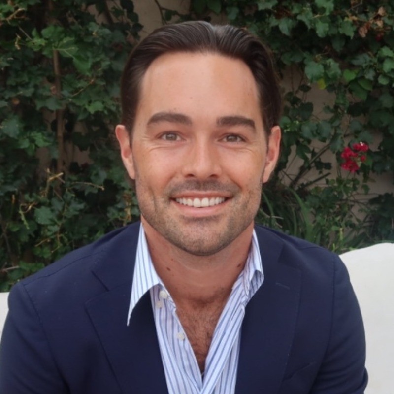 A man with dark hair wearing a navy blazer and striped shirt sits outdoors in front of green leafy plants, smiling at the camera.