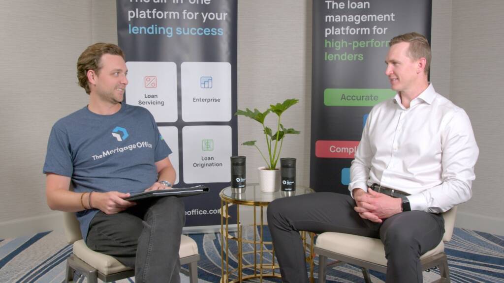 Two men sit facing each other in an office setting, having a conversation. Behind them are banners promoting lending software platforms. A small table with a plant and cups is between them.