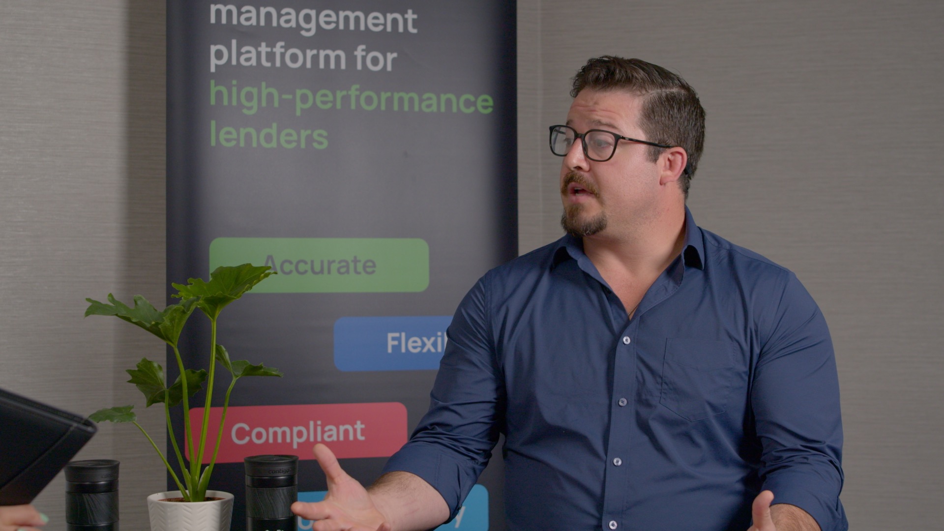 A man in a blue shirt and glasses speaks while gesturing with his hands, seated beside a plant; a business banner is visible in the background.