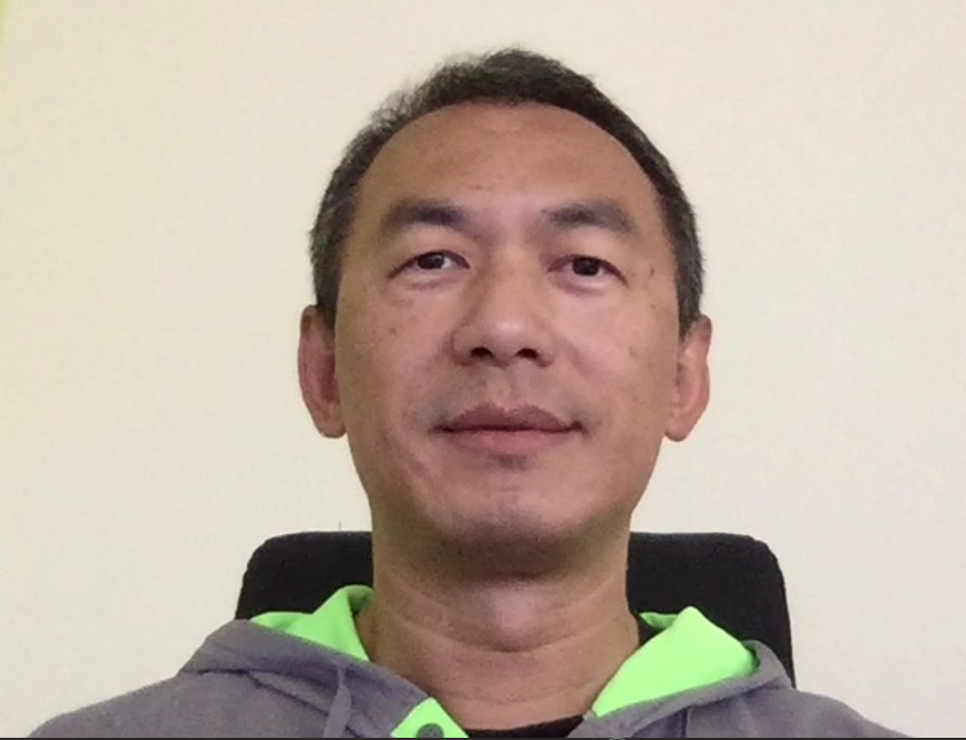 A man with short dark hair is wearing a gray hoodie with a neon green lining, sitting against a plain light-colored wall.