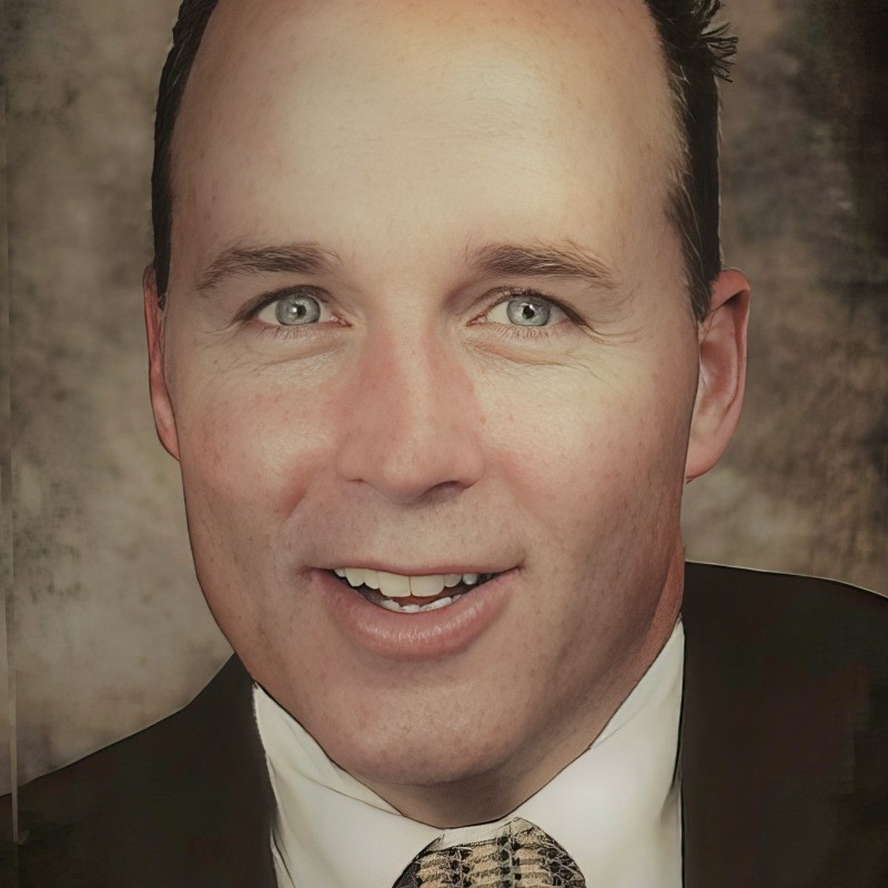 A man in a suit and patterned tie smiles at the camera against a brown, textured background.