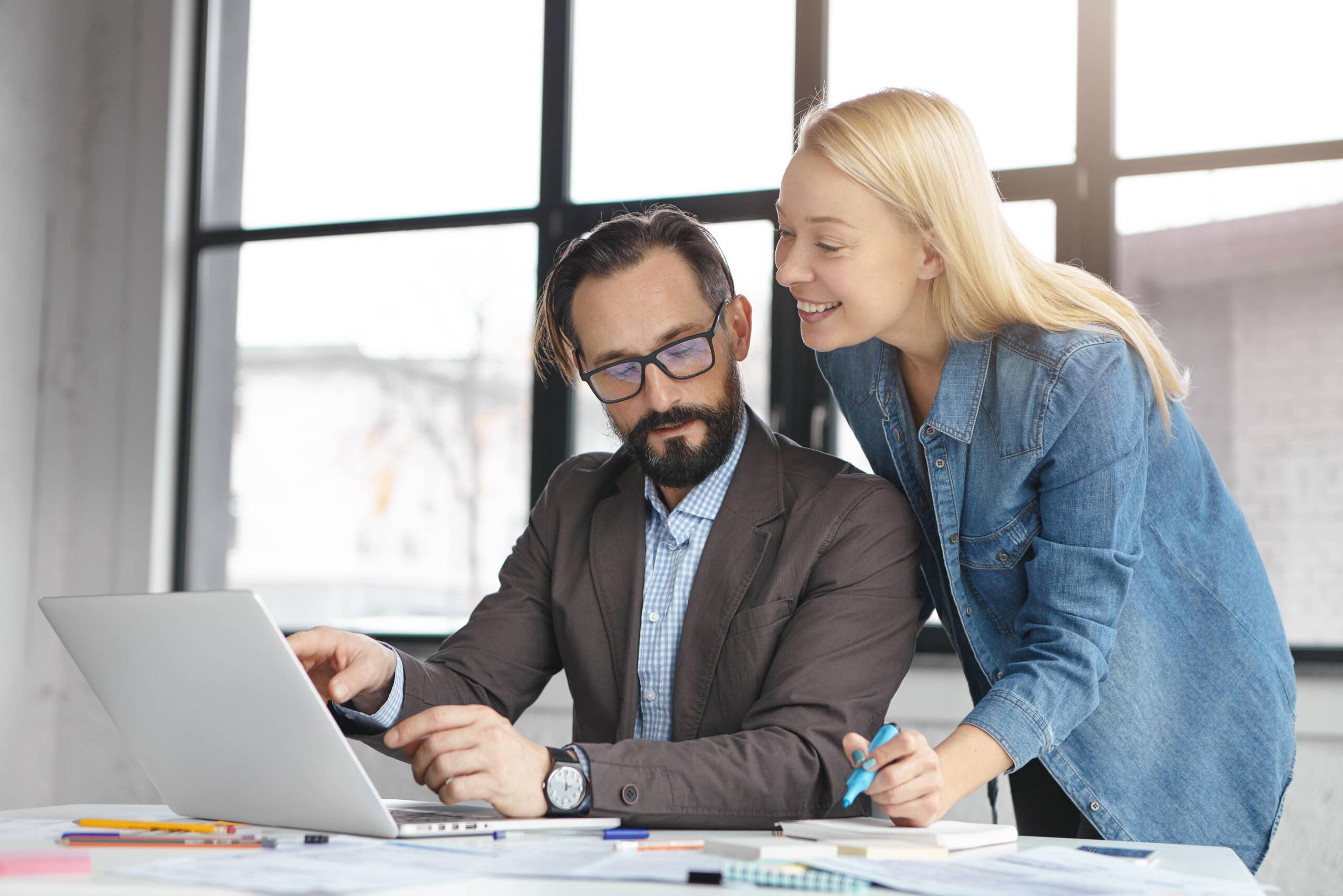 A man in a suit works on a laptop while a woman in a denim shirt stands beside him, smiling and holding a notepad and pen, in a bright office setting.