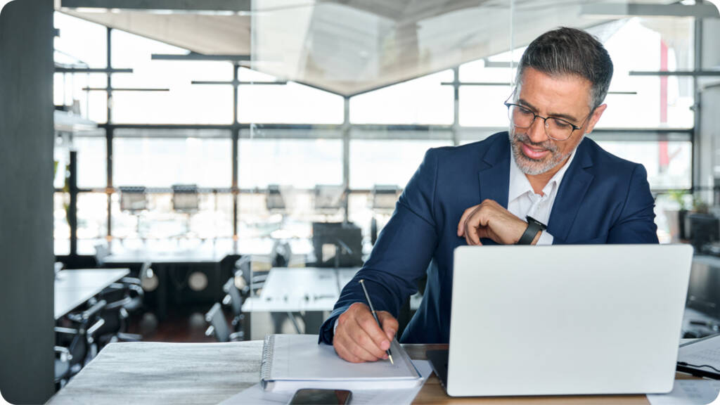 A man in a suit works at a desk with a laptop, takes notes in a notebook, and checks his smartwatch in a modern office setting.