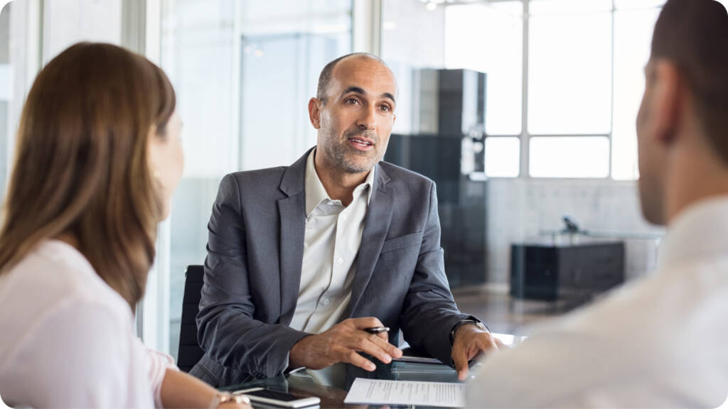 A man in a suit talks to two people across a desk in a modern office, holding a pen and paper.