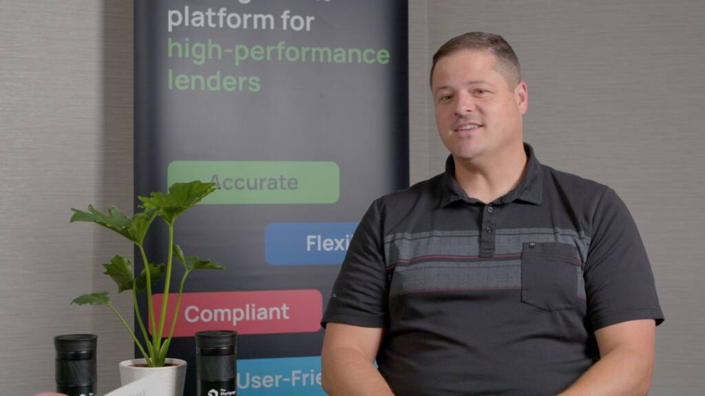 A man in a black polo shirt sits indoors next to a potted plant, with a banner about high-performance lenders in the background.