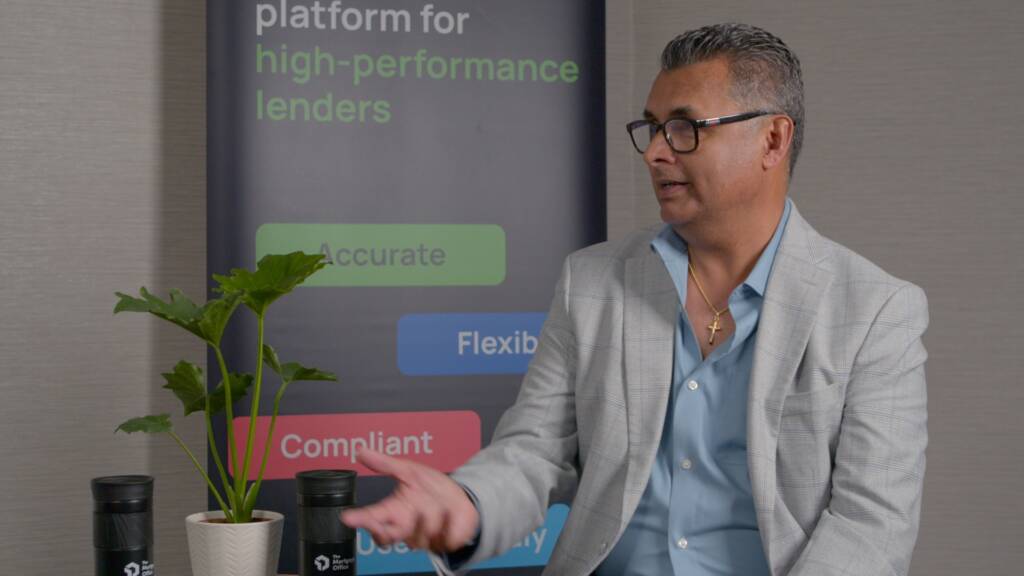 A man in a light gray suit jacket speaks while seated next to a potted plant, with a business banner in the background displaying keywords like "Accurate" and "Flexible.