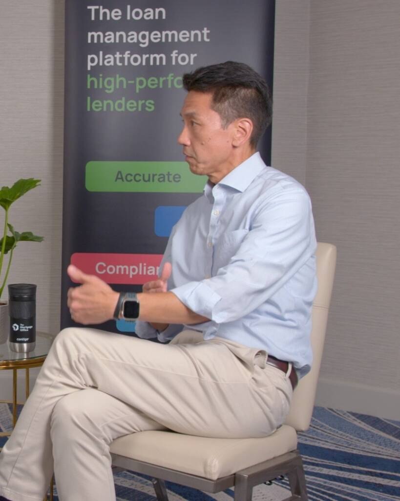 A man in a light blue shirt and beige pants sits cross-legged, gesturing with his hands, with a promotional banner about loan management software in the background.