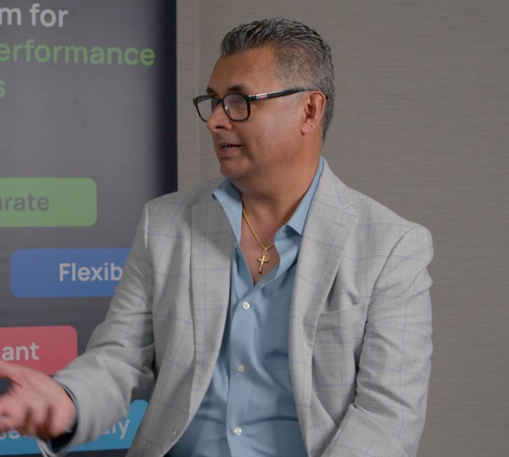 A man in a light gray blazer and blue shirt sits in front of a banner, gesturing with his hand. He wears glasses and a gold necklace with a cross.