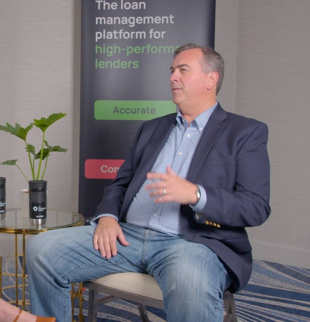 A man in a blazer and jeans sits on a chair in front of a banner about loan management platforms, gesturing with his right hand.