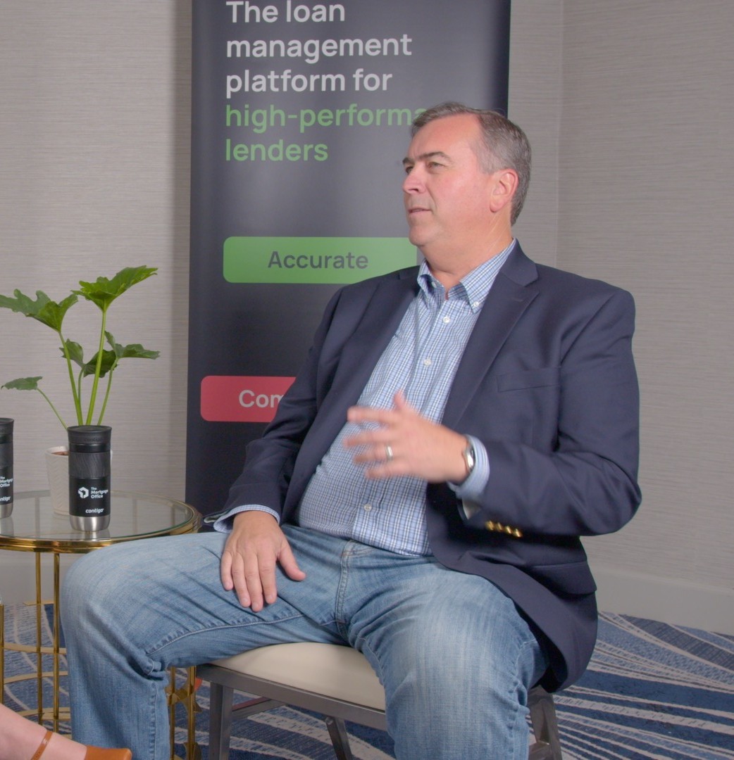 A man in a blazer and jeans sits on a chair in front of a banner about loan management platforms, gesturing with his right hand.