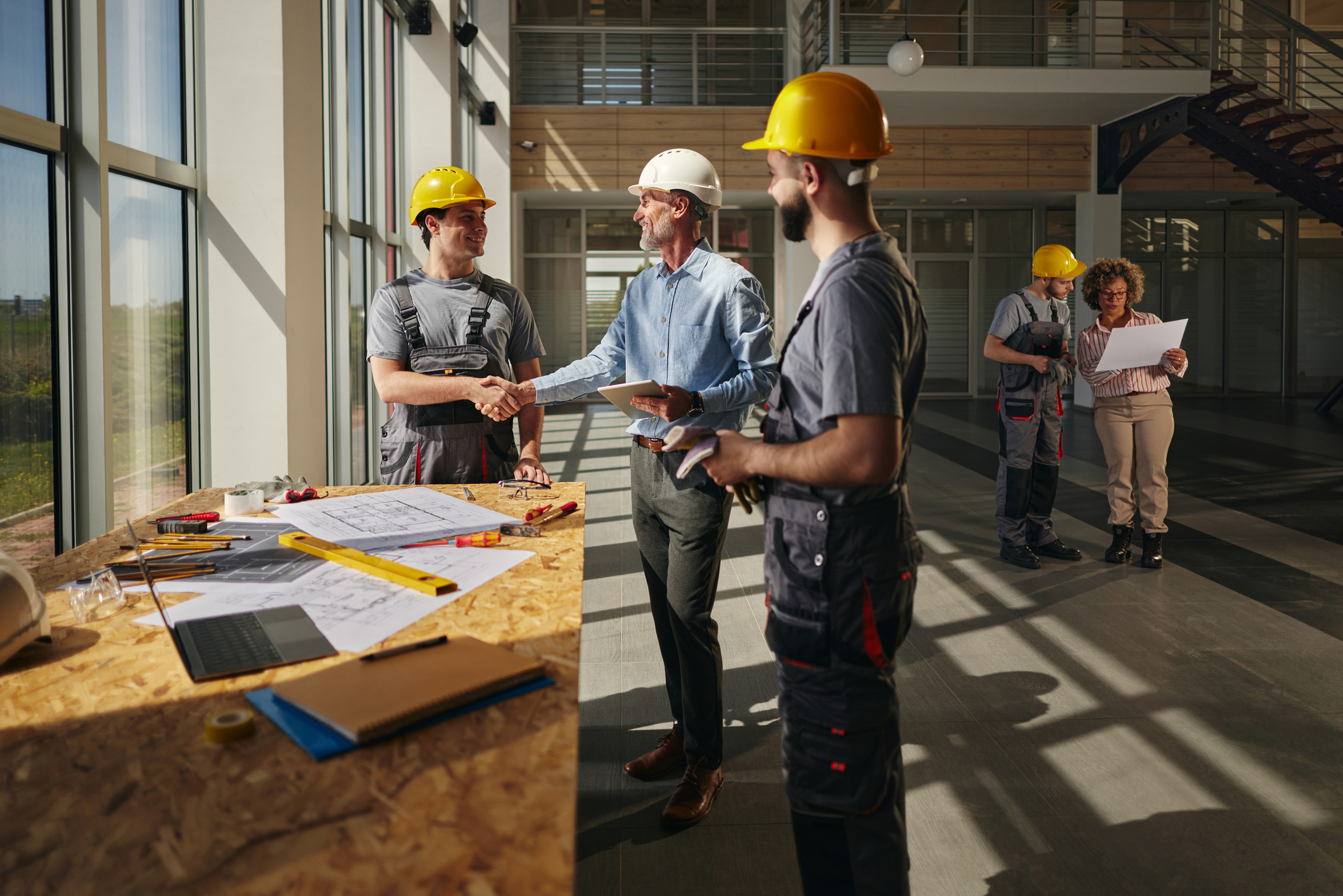 Two construction workers in hard hats shake hands with a man in business attire by a table with blueprints, while others review plans in the background of a bright building interior.