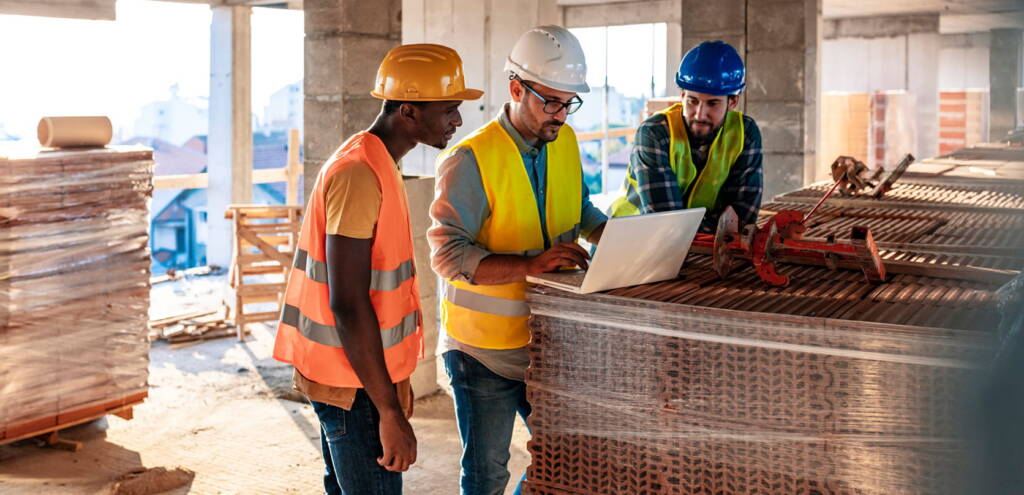 Three construction workers wearing safety vests and helmets review information on a laptop at a construction site, surrounded by building materials.