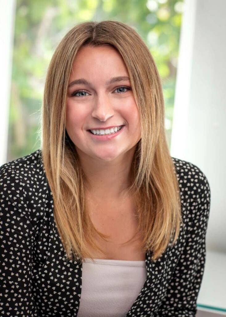 A woman with straight blonde hair wearing a patterned black jacket over a white top smiles while sitting indoors in front of a blurred background.