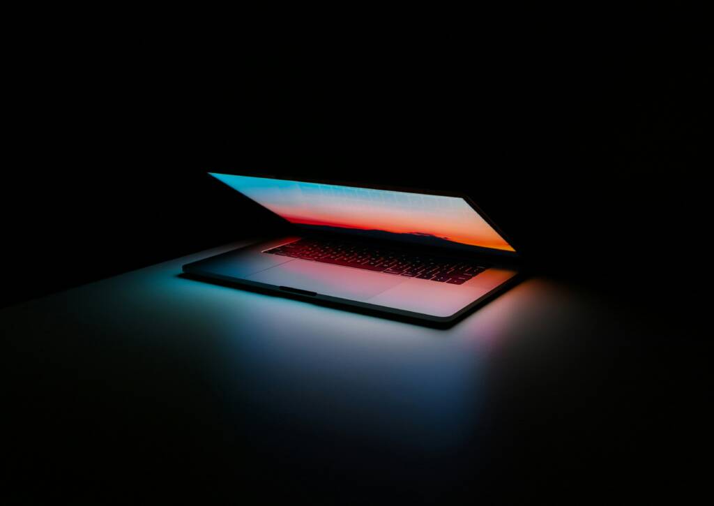 A partially closed laptop sits on a dark surface, its screen displaying a colorful gradient that reflects onto the table.