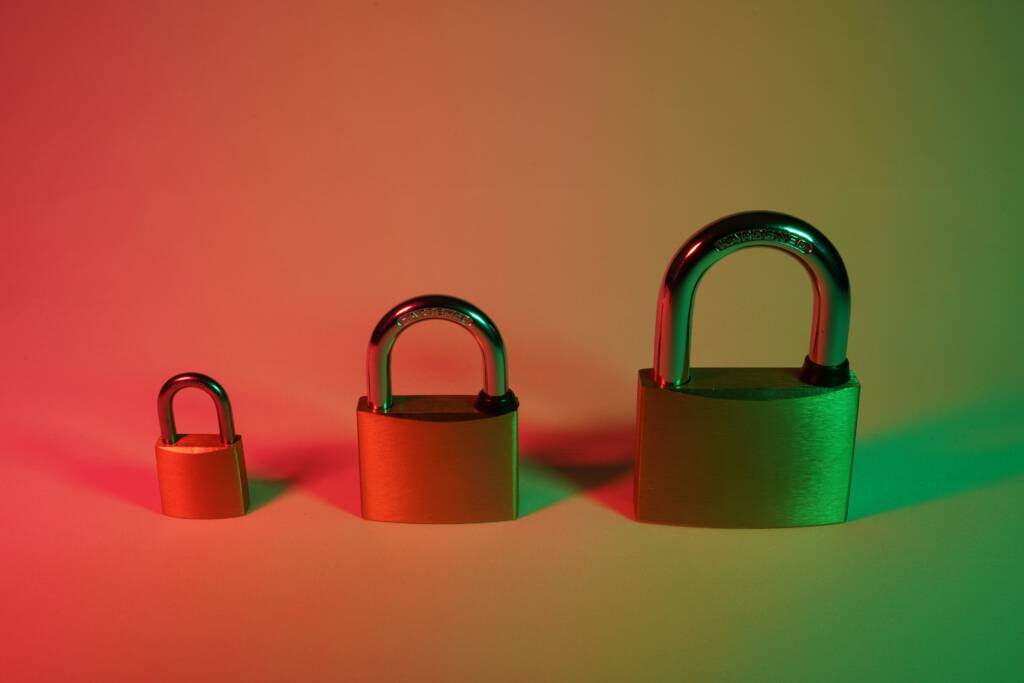 Three brass padlocks of varying sizes are arranged in a row from smallest to largest on a surface with red and green lighting.