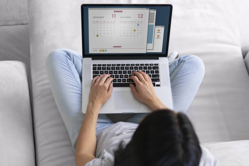 Person sitting on a couch using a laptop with a calendar displayed on the screen, viewed from above.